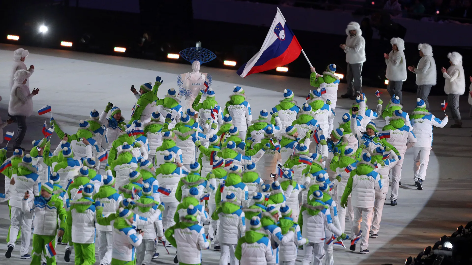 Slovenska ekipa nosi zastavo in se oblači v bele in zelene jakne ter modre kape, medtem ko paradejo na prireditvi.