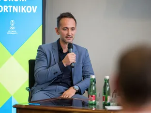 Moški v sivi obleki drži mikrofon in govori, medtem ko so na mizi steklenice vode.