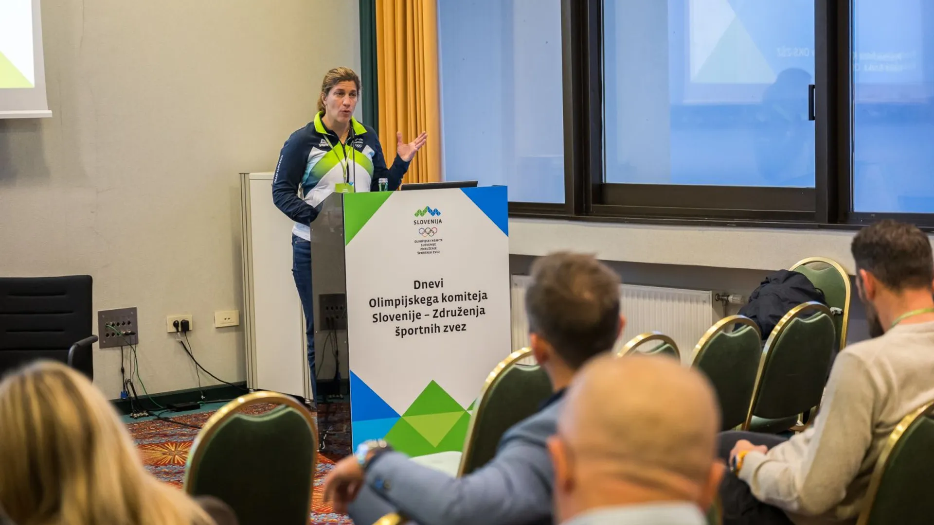 Ženska v športni obleki govori za mizo z napisom "Dnevi Olimpijskega komiteja Slovenije - Združenja športnih zvez".