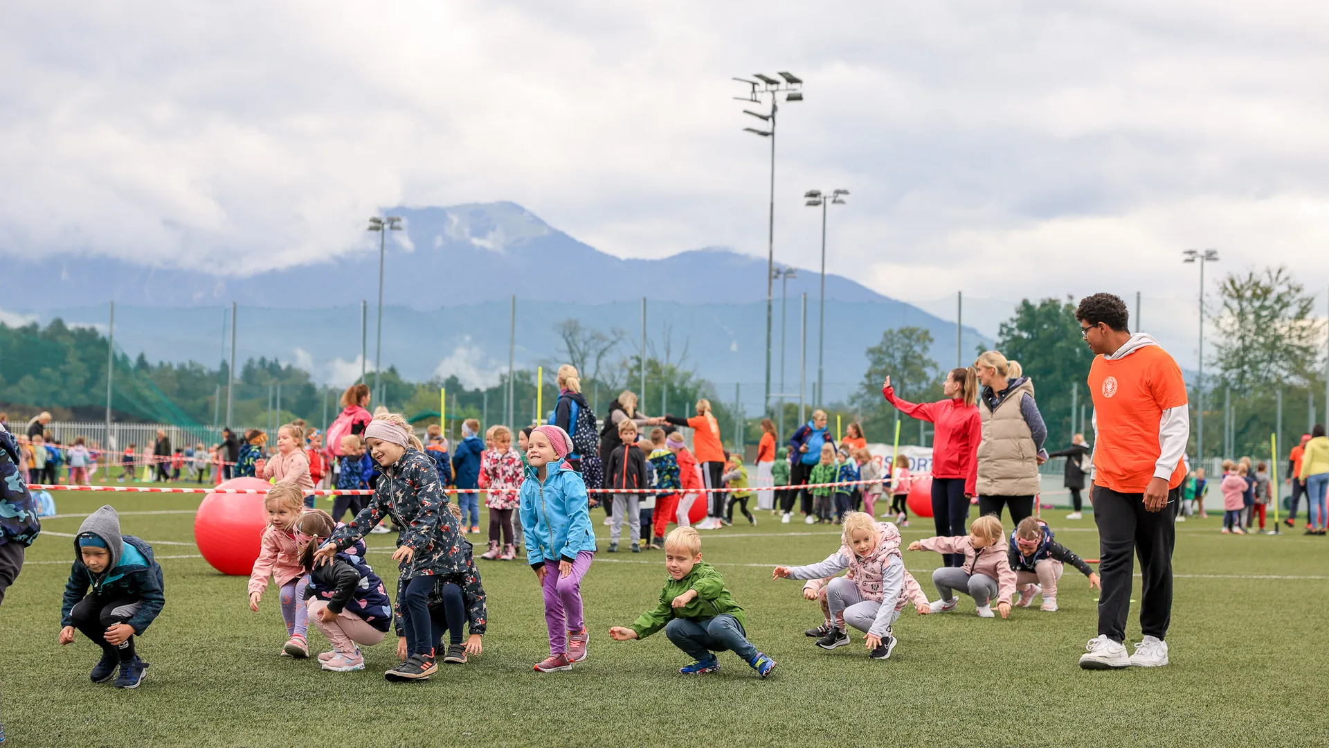 Otroci se igrajo na travnati površini, nekateri se sklanjajo, drugi stojijo, oblečeni so v različne barvite jakne.