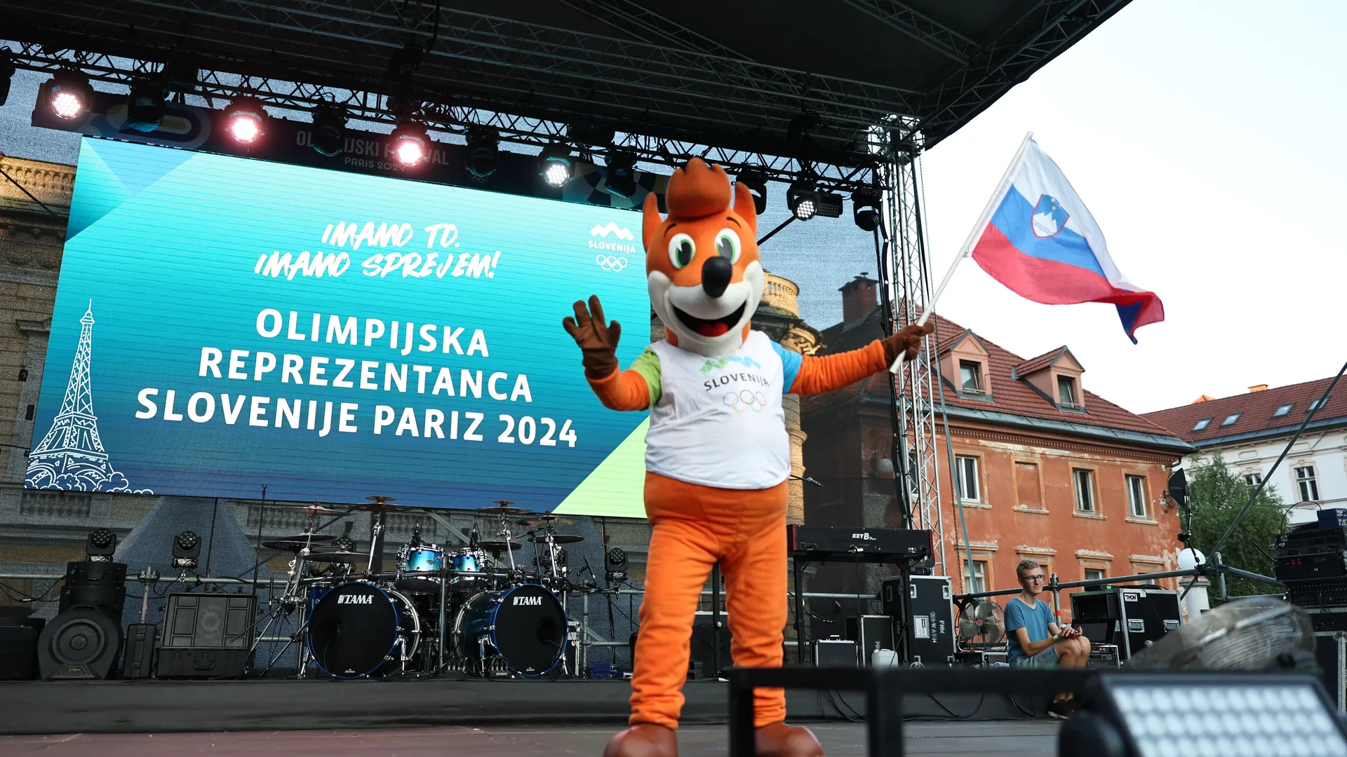 Maskota Foksi v oranžni obleki drži slovensko zastavo, medtem ko je na ozadju zaslon z napisom "OLIMPIJSKA REPREZENTANCA SLOVENIJE PARIZ 2024.