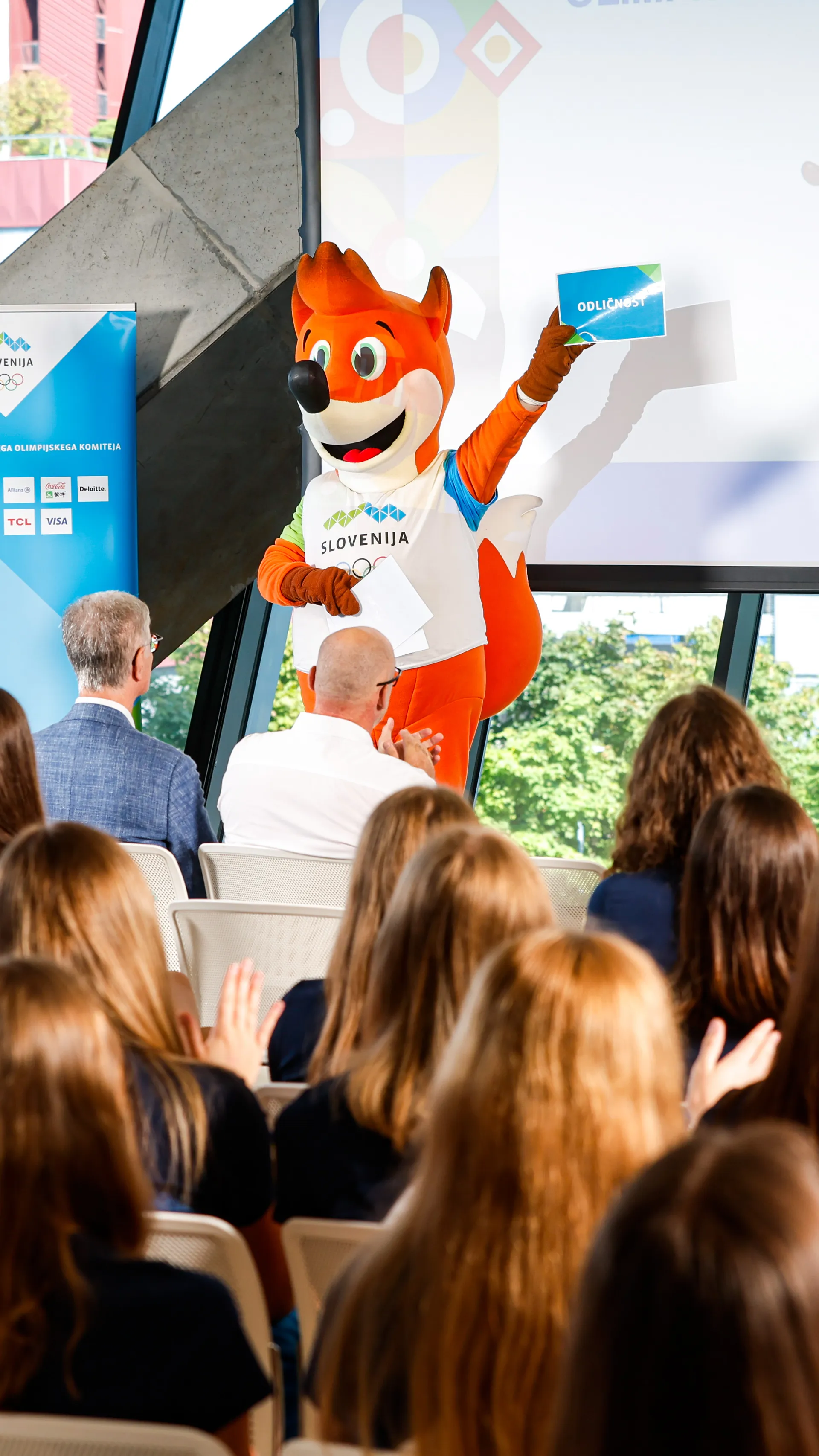 Maskota v oranžni obleki drži znak, medtem ko športnica v modri obleki bere papirje pred občinstvom.