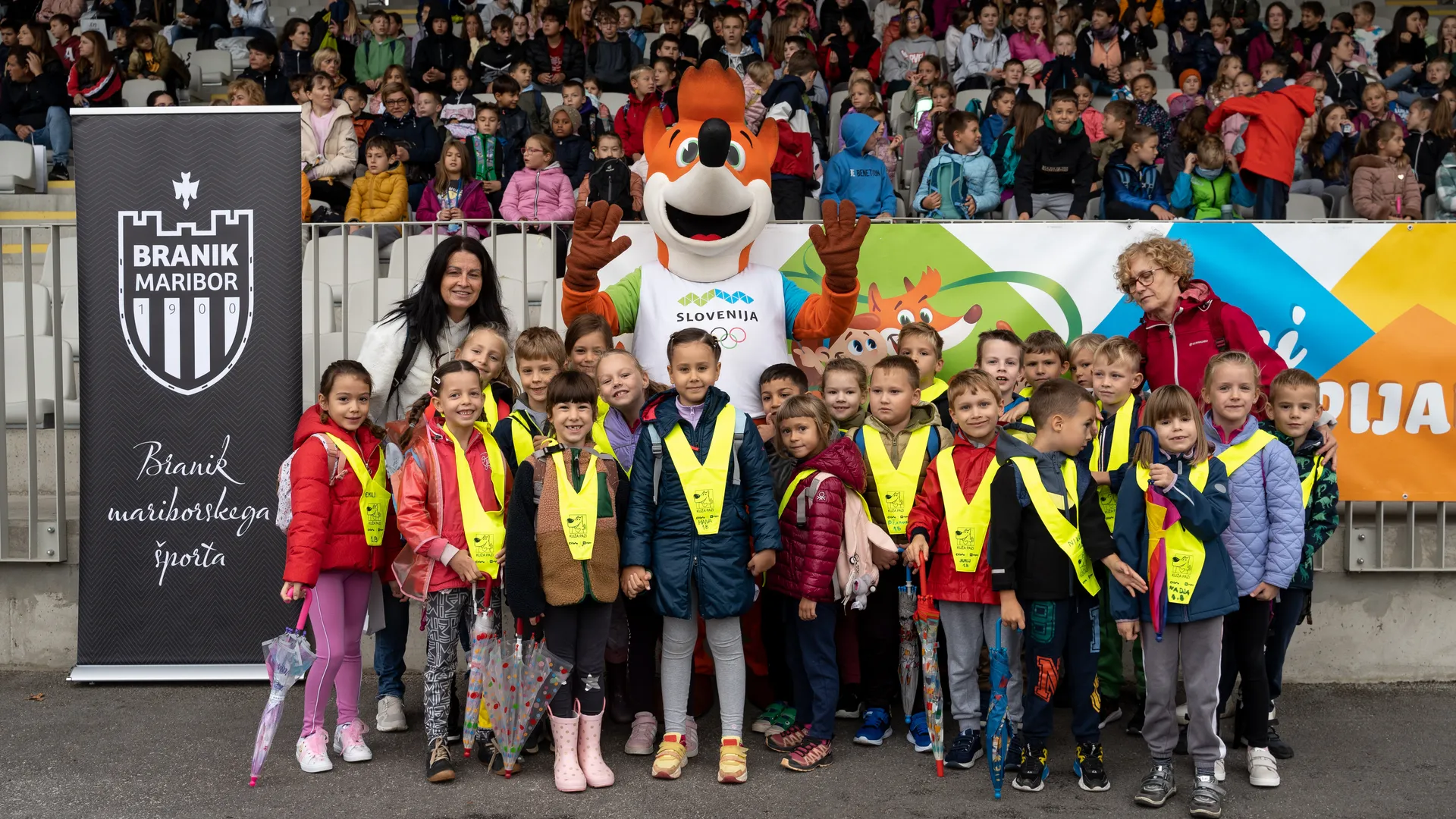Skupina otrok v rumenih jopičih stoji pred maskoto in transparentom z napisom "Slovenija" ter logotipom "Branik Maribor".