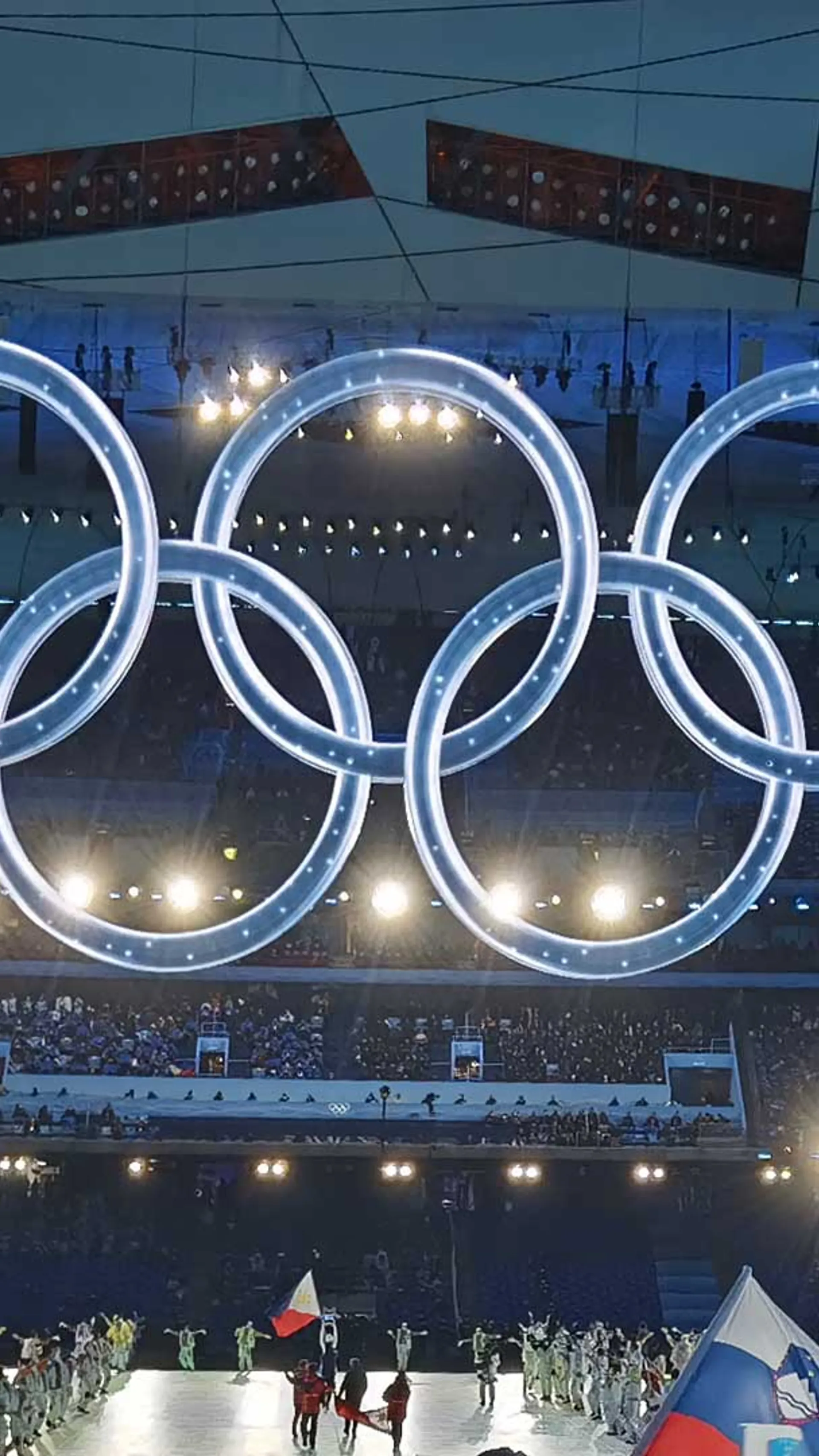 Na slovesnosti so prikazani olimpijski krogi nad množico ljudi, ki nosijo zastave in se udeležujejo dogodka.