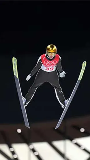 Smučar s številko 39 in zlato čelado skače v zrak z razpršenimi smučmi.