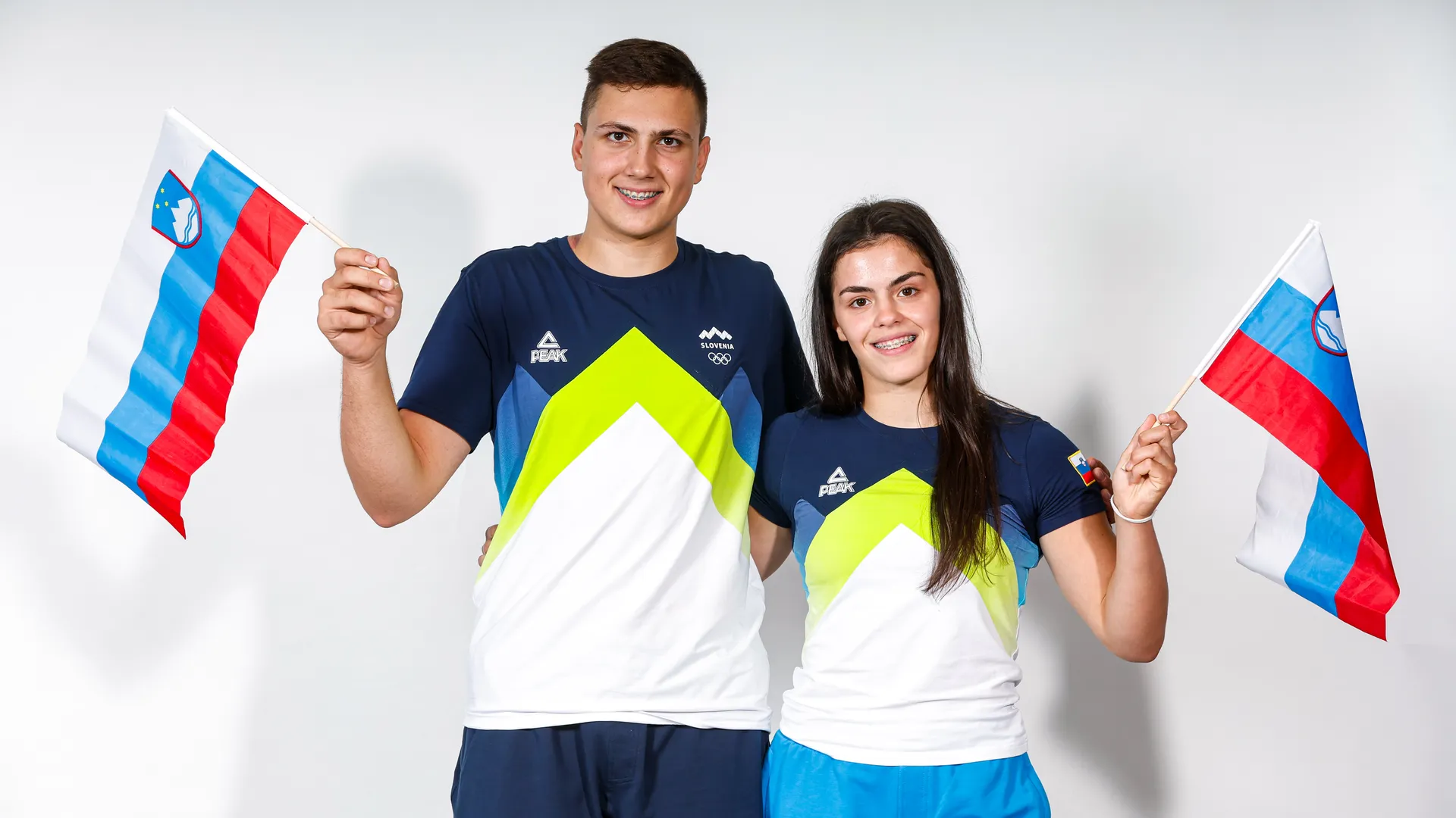 Moški in ženska v športnih majicah z zelenimi in modrimi detajli, vsak drži slovensko zastavo.